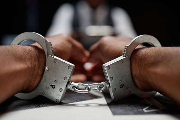 Close up of hands in handcuffs on a desk with man sitting opposite